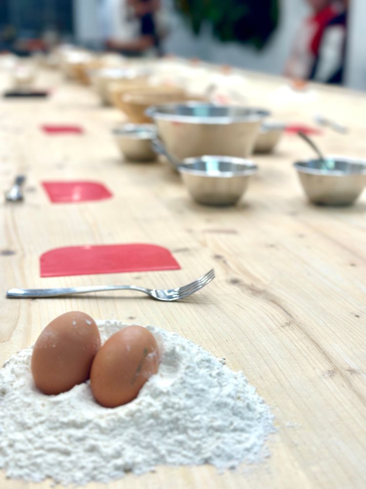 Two eggs sit in a pile of 00 flour on a long wooden table, with a fork and red bench scraper just beyond 