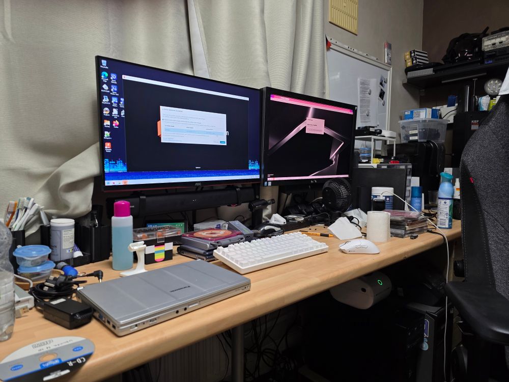 A photo of my workbench. On it are various tools, a Panasonic Let's Note, a VAIO C1MGP, some discs, and on the two displays mounted to monitor arms is my workbench PC's desktop running Fusion.