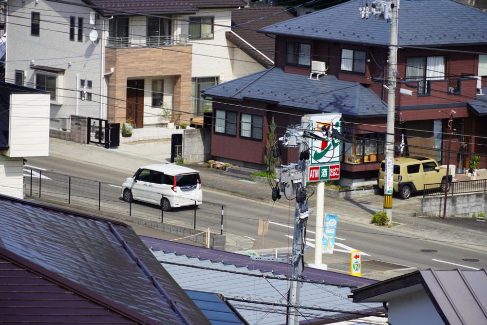 A zoomed in shot of a Japanese street. Power lines running everywhere. A Daihatsu Thor (I think?) drives by the 7-Eleven.