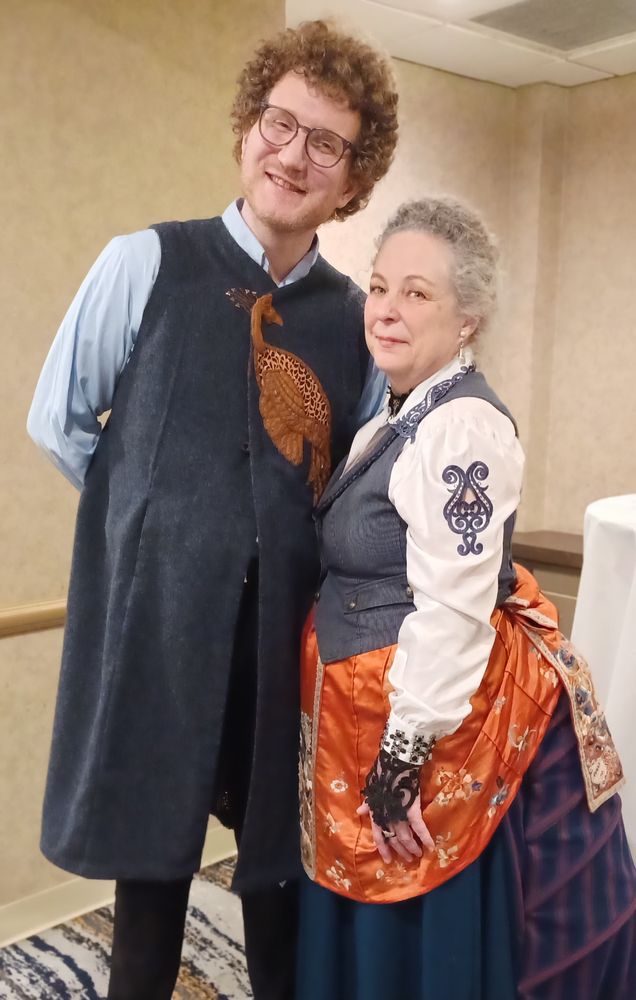 At left is my Peacock Vest being modeled by a male colleague.  It's a knee length navy wool vest, appliqued with a peacock in rust colored suedecloth both front and back. The peacock was inspired by the Peacock Doors at the Palmer House in Chicago, made from cast bronze by Tiffany in the 1880s. At right, I am wearing a Neo-Victorian ensemble made from a dark blue skirt, a gifted bustle in navy and wine striped denim, a rust colored silk Chinese skirt (too damaged to restore so I crafted it into a drape and bustle).  The blouse was stark white until I tried to dye it, but only the embroidery on the collar and sleeves took the dye. My navy waistcoat is a thrift store find that I tricked out with some passementerie and new brass buttons.  All materials in both pieces were recycled from previously owned sources with the exception of the square studs in the peacock's tail at left, and the gifted bustle at right.