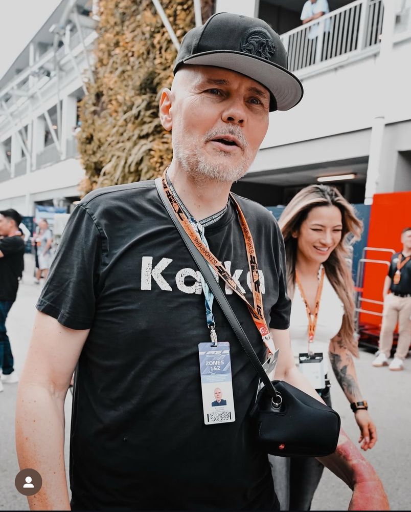 Cantor Billy corgan usando camisa preta que parece que tá escrito Kafka mas tem um monte de cordão de credencial tapando a letra do meio, usando boné preto, barba grisalha, andando pelo paddock