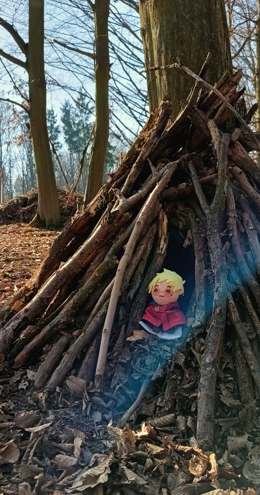 Photo of a TeruVa plushie sitting at the entrance of a hut made of branches in a forest. He is living the life in the woods we all crave so much.

