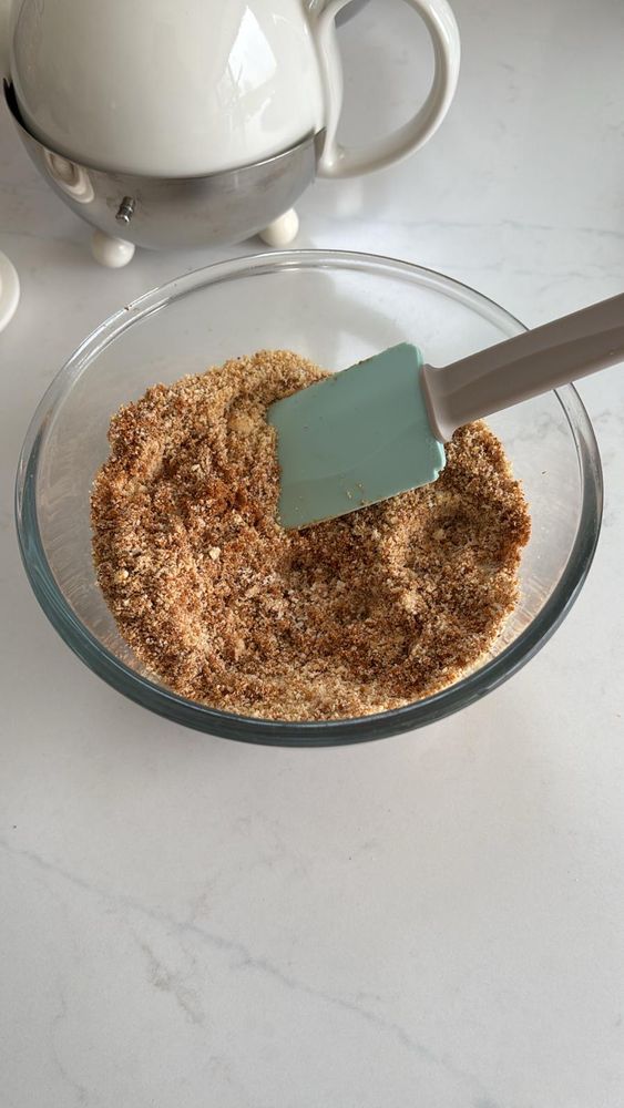 a glass bowl containing a mix of brown and cream toasted ground hazelnuts and almonds, with a blue and a grey plastic spatula sticking out the bowl