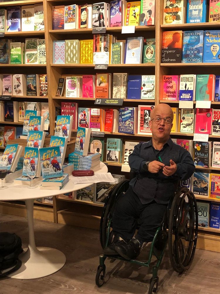 Tom Shakespeare, a bald man in a blue shirt and a wheelchair, talking about THE ENDS at this week's launch beside a display of the book, in front of bookshelves.