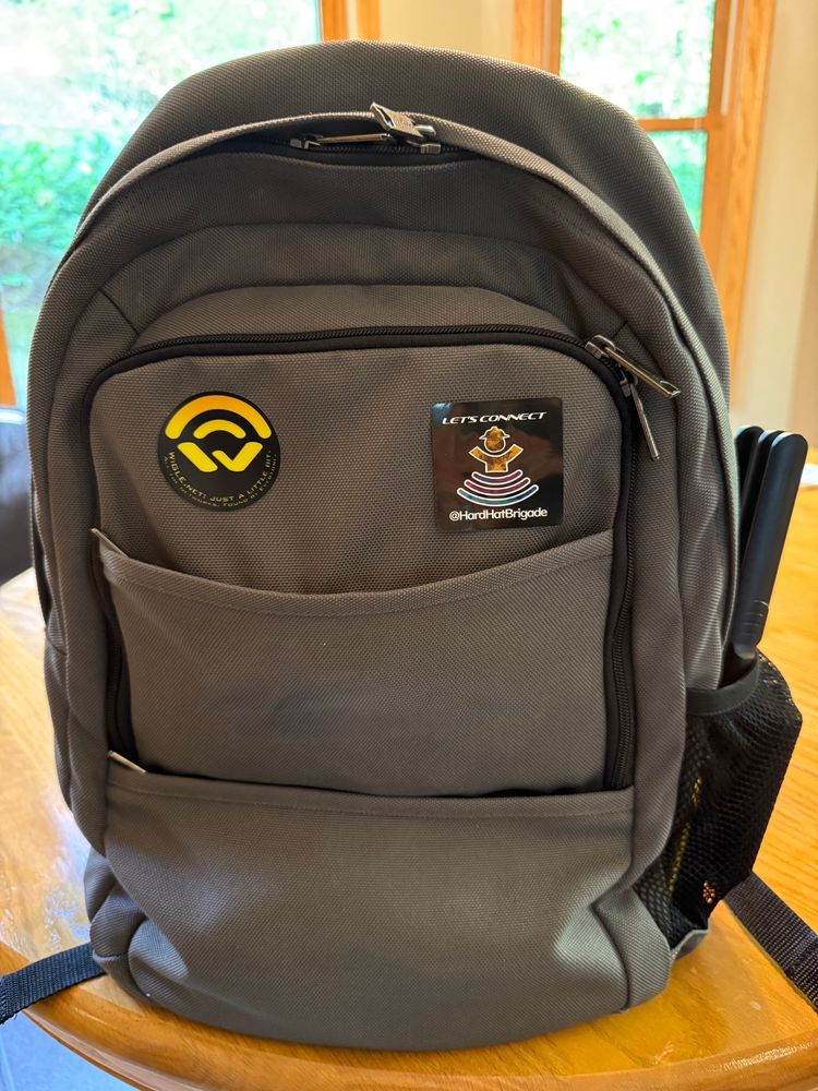 A gray backpack with yellow wigle.net sticker and a black Hardhat Brigade sticker. A wardriver is in the side pocket.