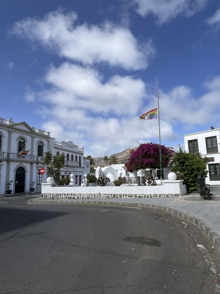 Eine Regenbogenflagge mit 6 Farben im Gemeindepark von Haría, Lanzarote. Das Gemeindehaus steht davor. 
