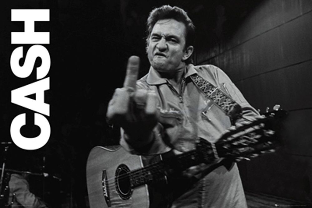 A black-and-white photograph shows a stern-faced Johnny Cash on stage at San Quentin prison in 1969, defiantly flipping the middle finger toward the camera. He is wearing his signature all-black outfit.