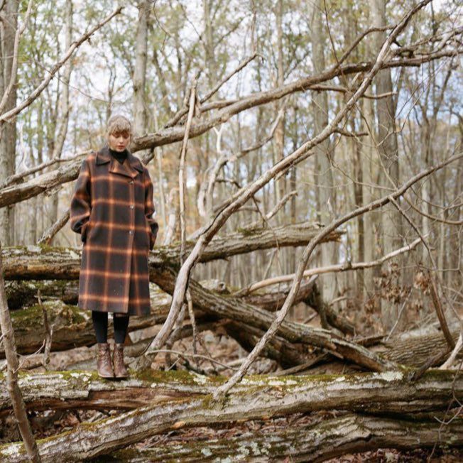 Taylor swift wearing a plaid coat. She's standing on a big tree branch