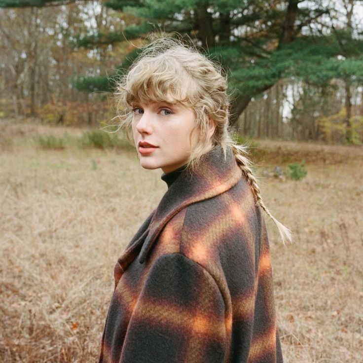 taylor swift wearing a plaid coat. She's looking back at the camera and has braided hair. there's a big tree behind her with green leaves
