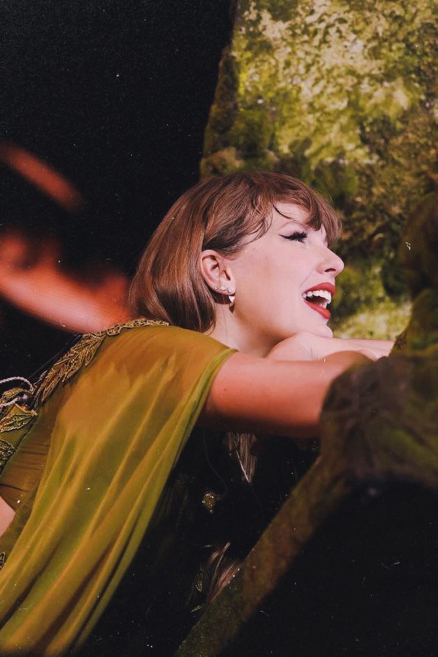taylor swift on stage at the eras tour, wearing the green folklore dresse. She's sitting at the piano and smiling