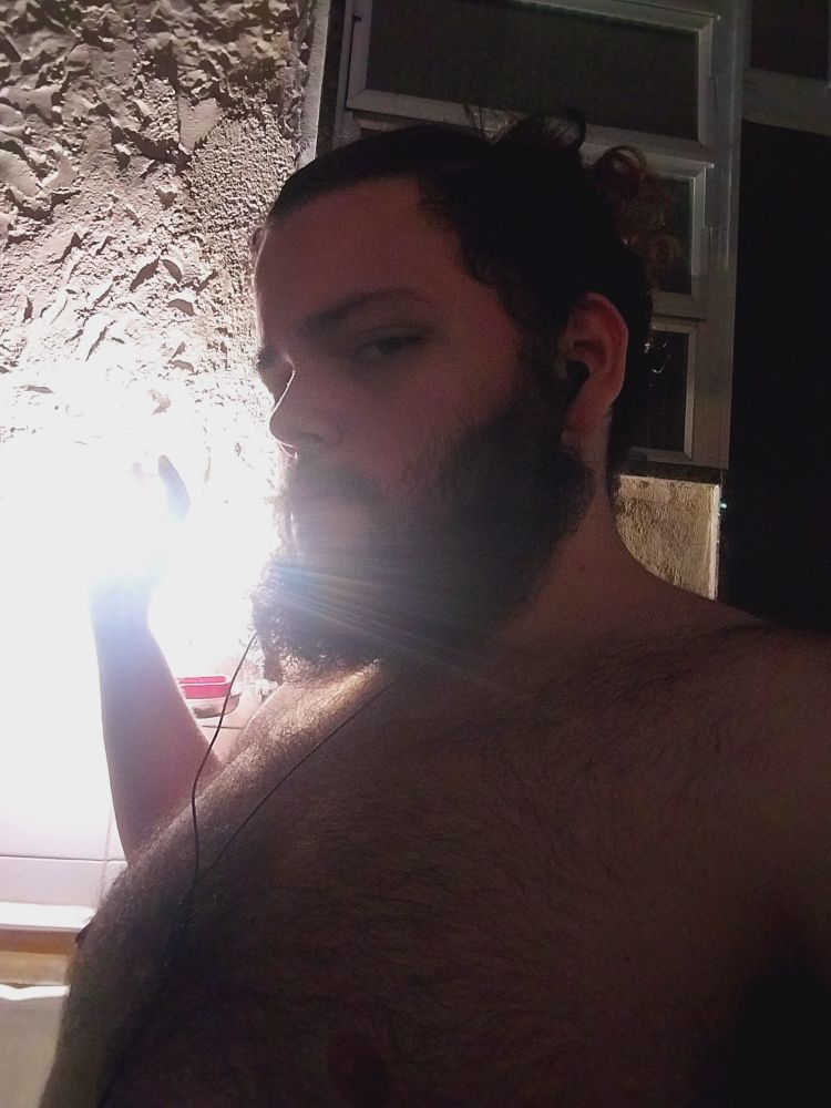 Homem barbudo de perfil, sem camisa com a mão levantada como se fosse pegar o foco de luz, foto tirada contra a luz