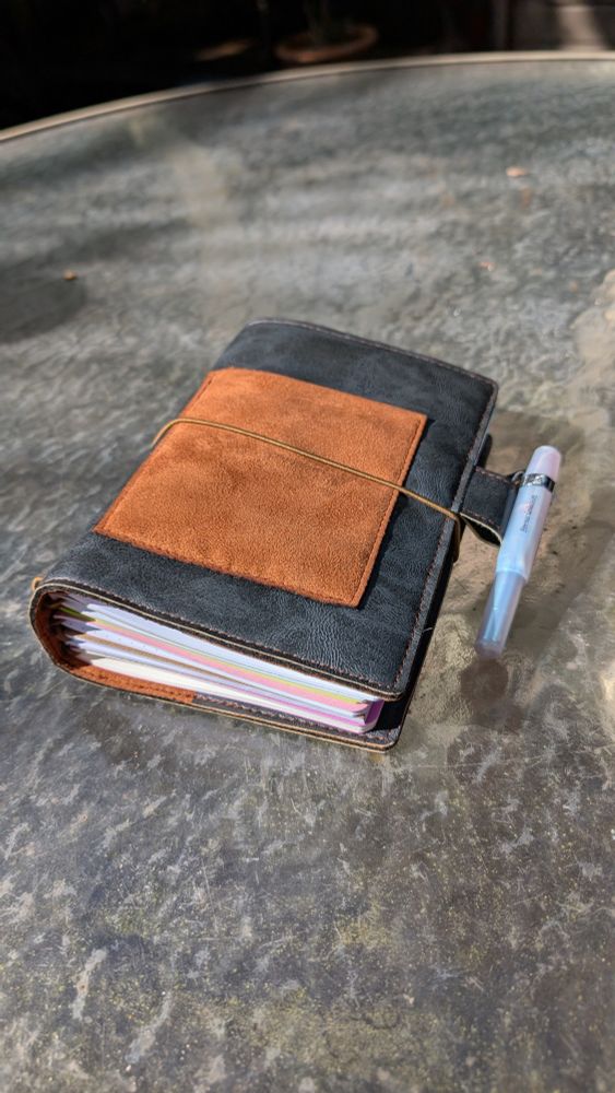 A closed black vegan leather traveler's journal with a brown pocket on the front is displayed on a glass table outside in natural light. It has a white pearlescent kaweco sport pen clipped to the pen clip and six notebook inserts are inside the journal cover.  