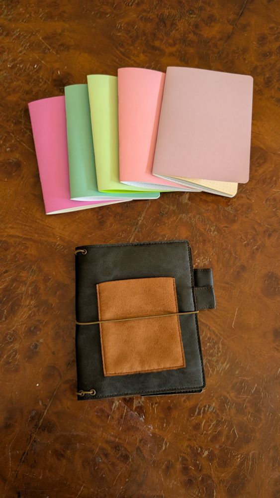Five pastel colored B6 size notebooks fanned out above a black vegan leather traveler's journal cover with a brown pocket on a wood table. 