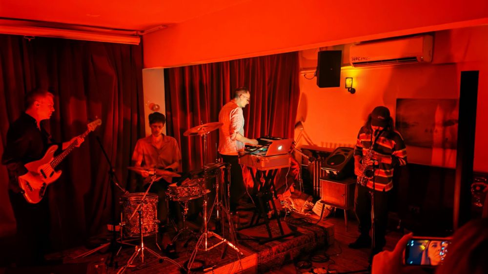 Kreidler, band on stage, from left Alex Paulick on bass, Thomas Klein on drums, Andreas Reihse on Synthesisers, with guest Timuçin Dündar on saxophone; room in red light