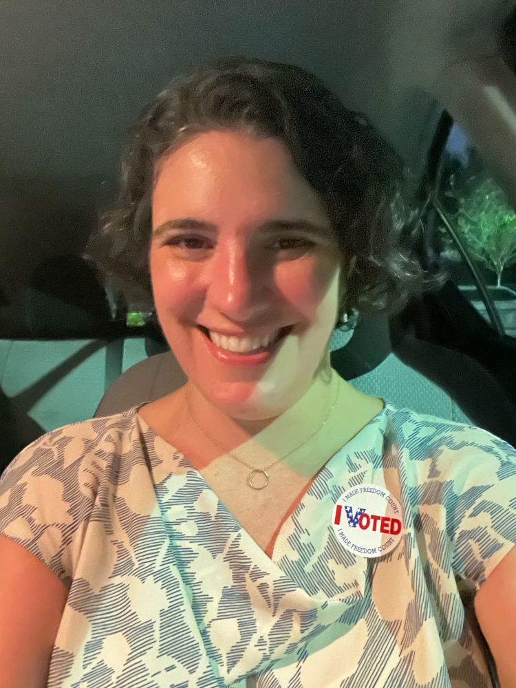 A picture of me, a middle-aged white woman, sitting in my car, smiling. I’m wearing a blue and white blouse and a sticker that says “I voted.”