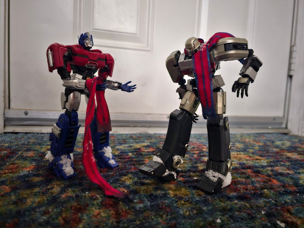 Orion and D16 standing on an entry rug, snow caught in their leg joints. Orion has removed his scarf 