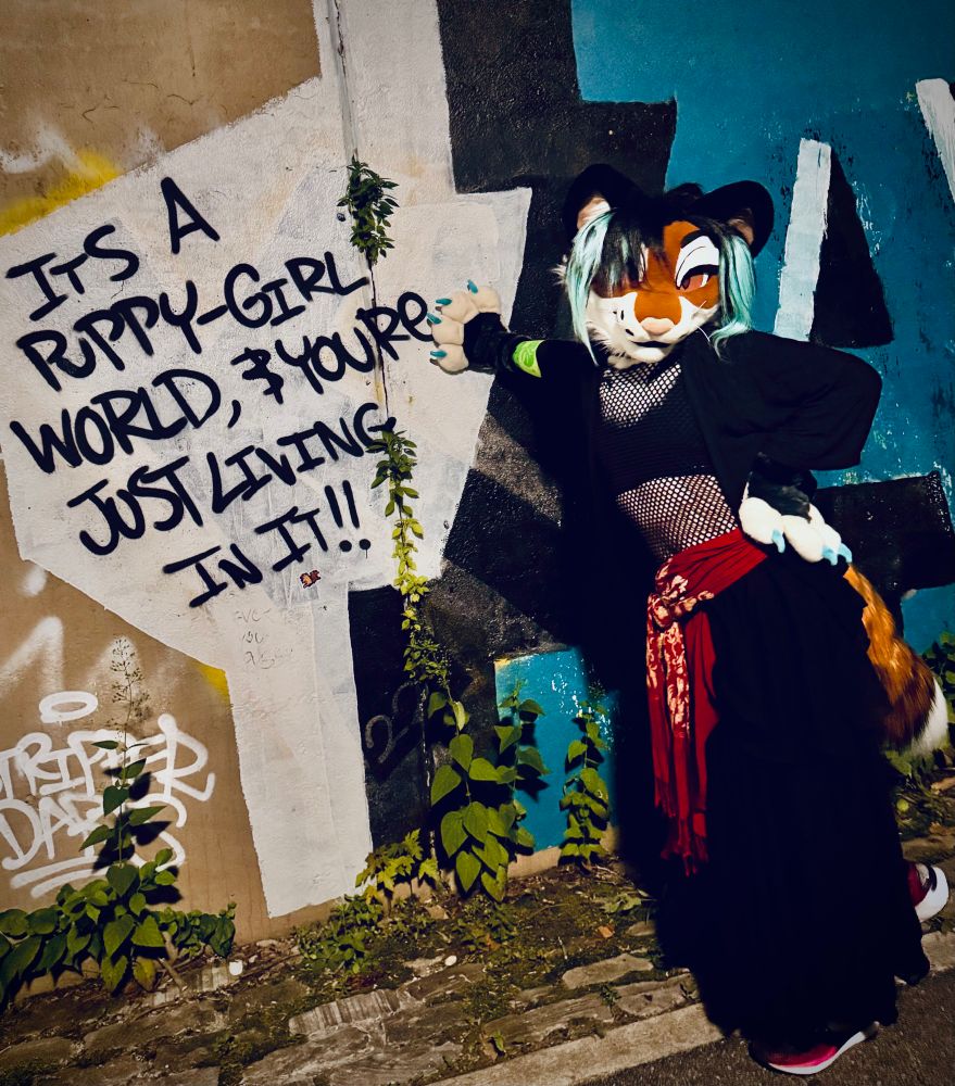 Talexis, a nonbinary tigerfox, dressed in a black and red outfit poses with a graffiti sign that reads, “It’s a puppy-girl world & you’re just living in it!!”