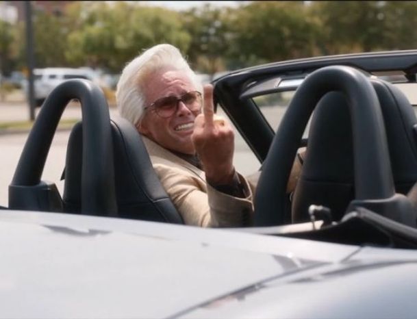 Uncle Baby Billy Freeman, an elderly gentleman with white hair, sunglasses, and prominent teeth, sitting in a convertible and spitefully, desperately flipping the middle finger over his shoulder