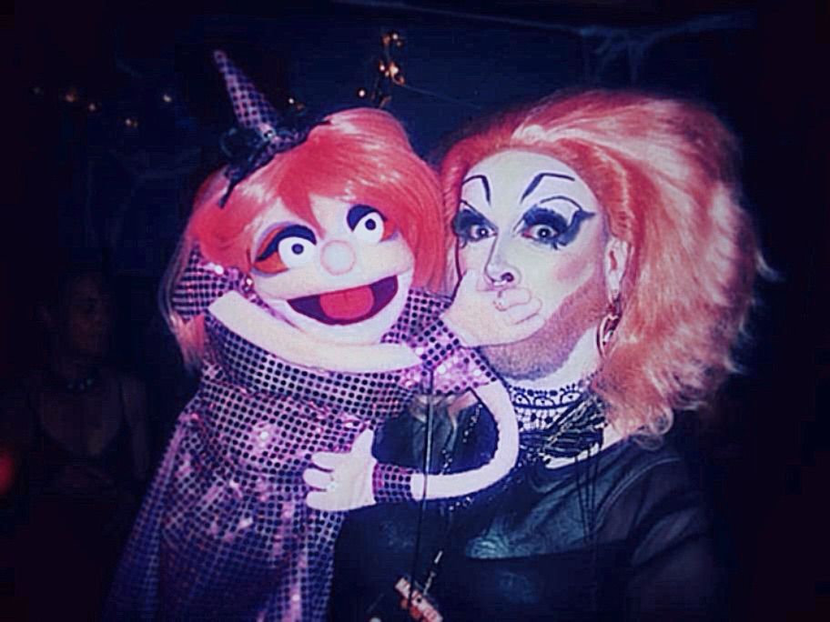 Eine Handpuppe die der Dragqueen den Mund zuhält.