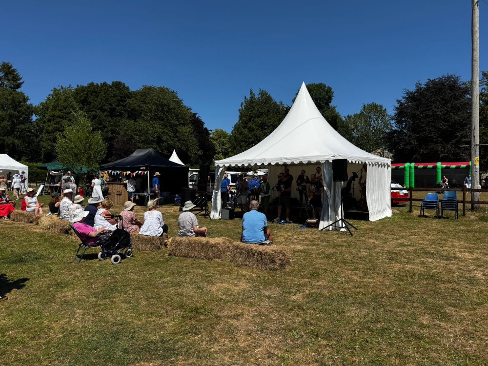 Villagers sat on hay bales watching the music in the tent - inflatables behind under flawless blue sky
