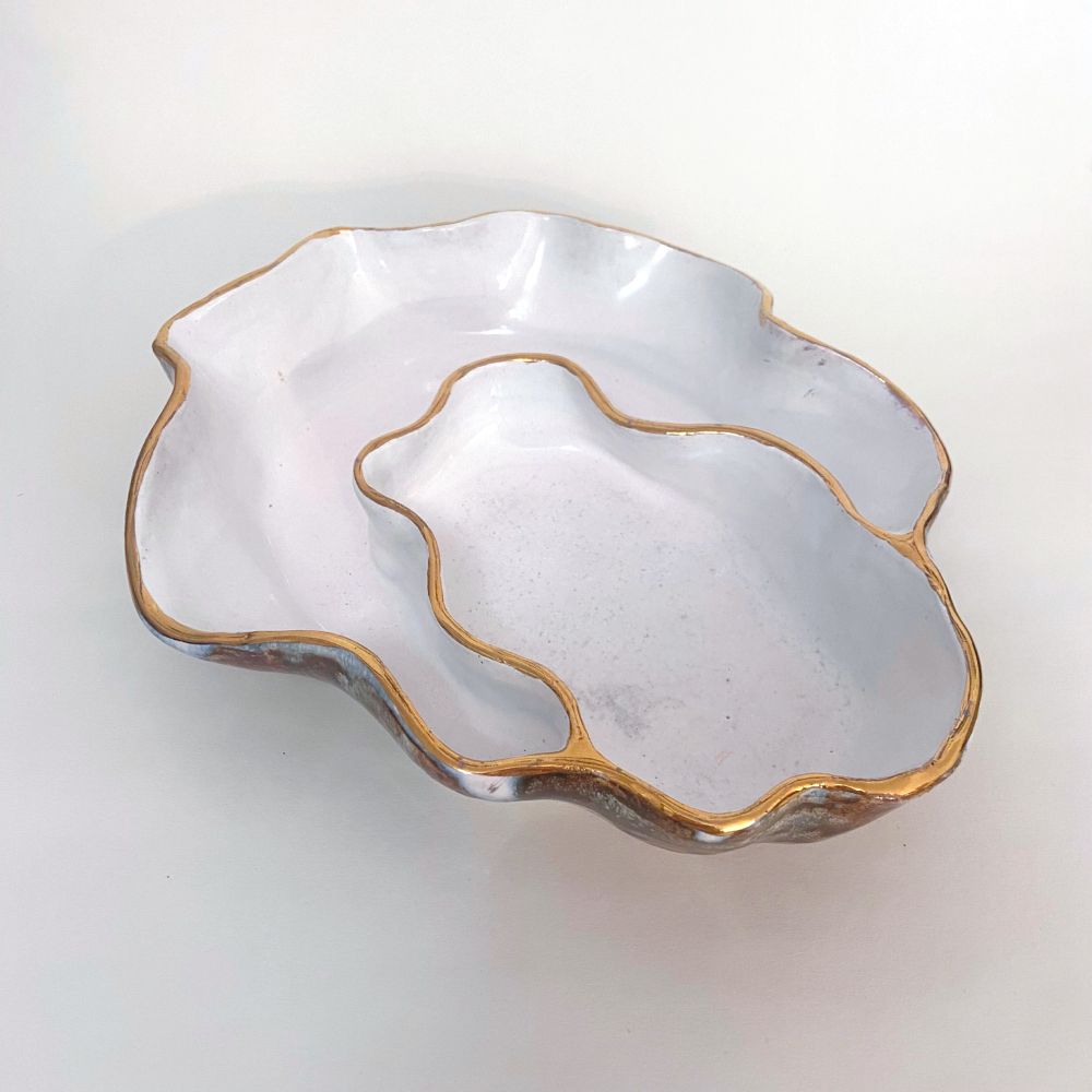 Three-quarter angle view of oyster-shaped bowl against a white background. The overall shape is like an oval or egg with a scalloped rim. There are two sections inside, one oval and one crescent shape. The rim has gold luster. The glaze inside is white, and the glaze outside is drippy gray and black.