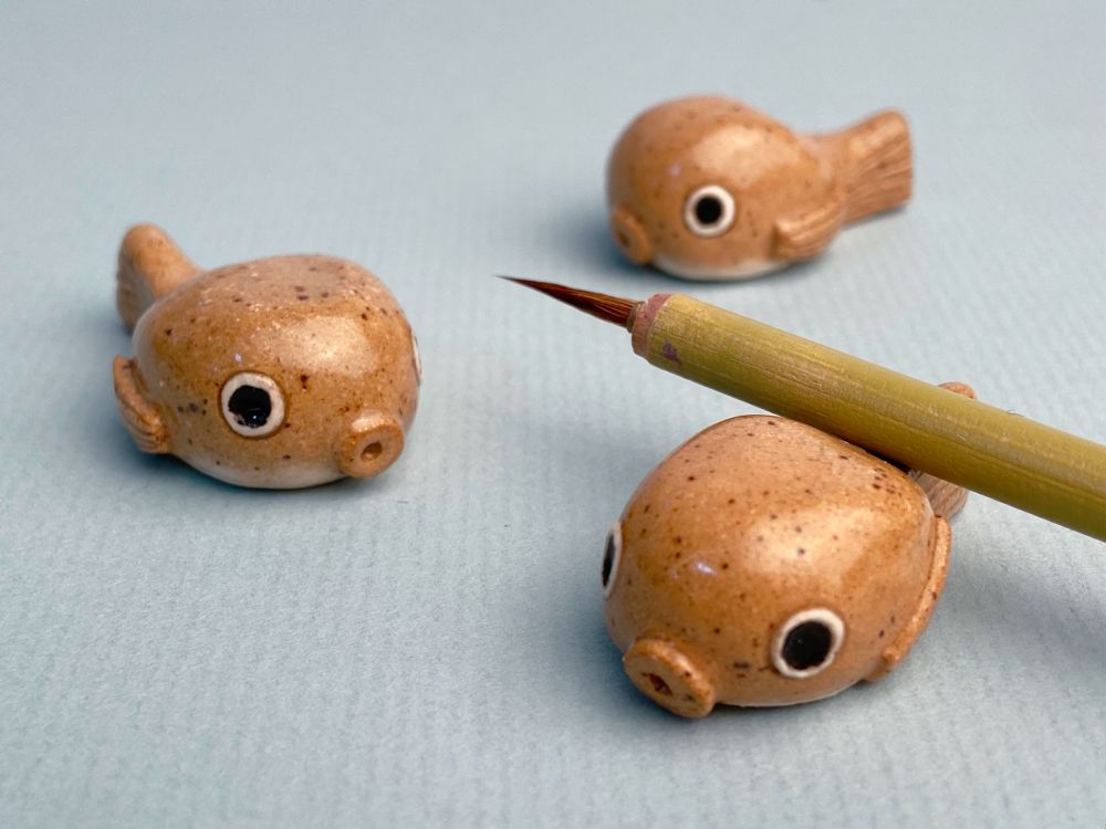 Three small ceramic figures of fugu (blowfish) on a blue-gray piece of paper. The frontmost one has a paintbrush resting on the narrowest part of the body, right before the tail. They are made out of a tan speckled clay and have big round cartoony eyes