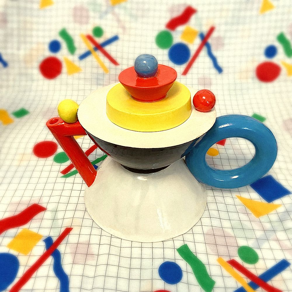Ceramic teapot against an 80s party tablecloth which is white with a black grid and red, yellow, blue, and green geometric shapes. The body of the teapot is hourglass-shaped, with the faces of the shape alternating between white and black. The handle is a blue torus/doughnut shape, and the spout is a red triangle shape. The lid is made up of a yellow cylinder red cone, and blue sphere. There is also a red sphere near the handle and a yellow sphere on the spout.