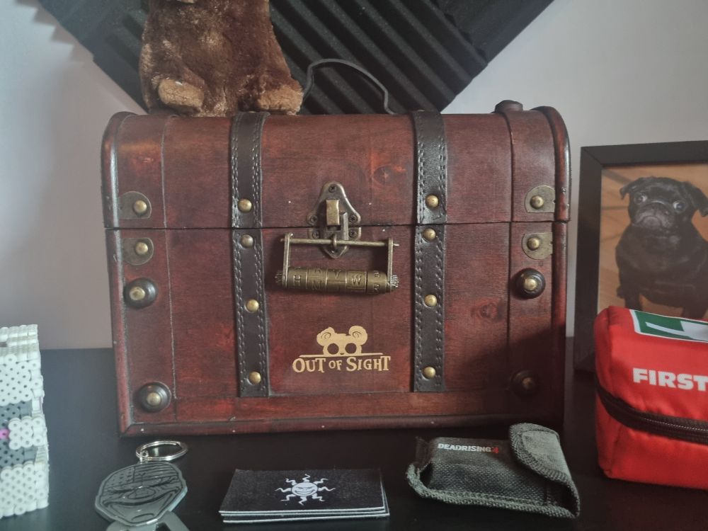 A wooden chest with leather details and a puzzle lock. The Out of Sight game logo can be seen engraved on the front of the box. Other game collectibles are surrounding it.