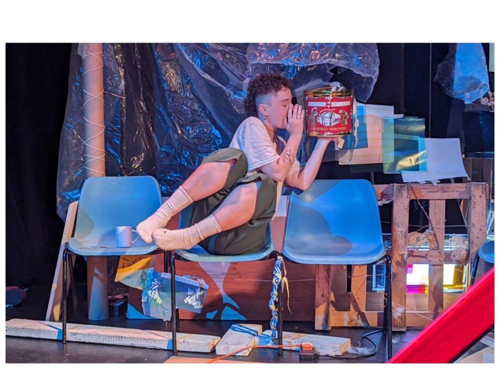 Photo of scamp sitting on a row of blue chairs. They are whispering into the side of a big can 