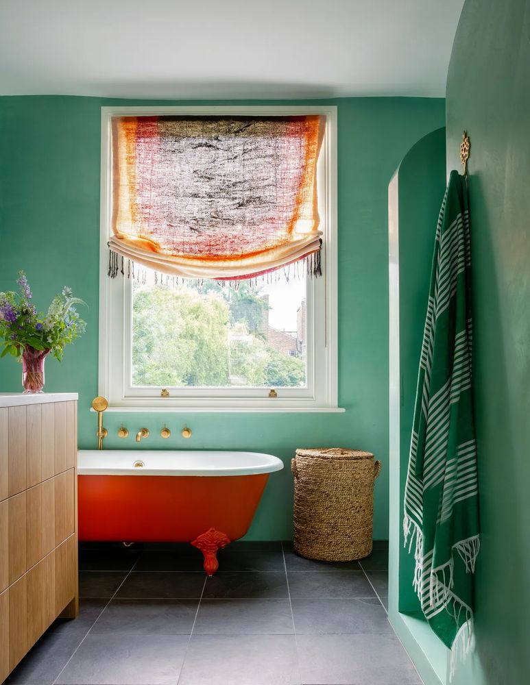 Red clawfoot bathtub, green tadelakt walls, Honor Devereux design for a South London Victorian home, custom Moroccan architectural arch shower stall, slate floor, hand woven basket, gold hardware faucet, custom window treatment inspired it all - a fringed throw turned into a semi-open woven scrim. House and Garden UK March 2024