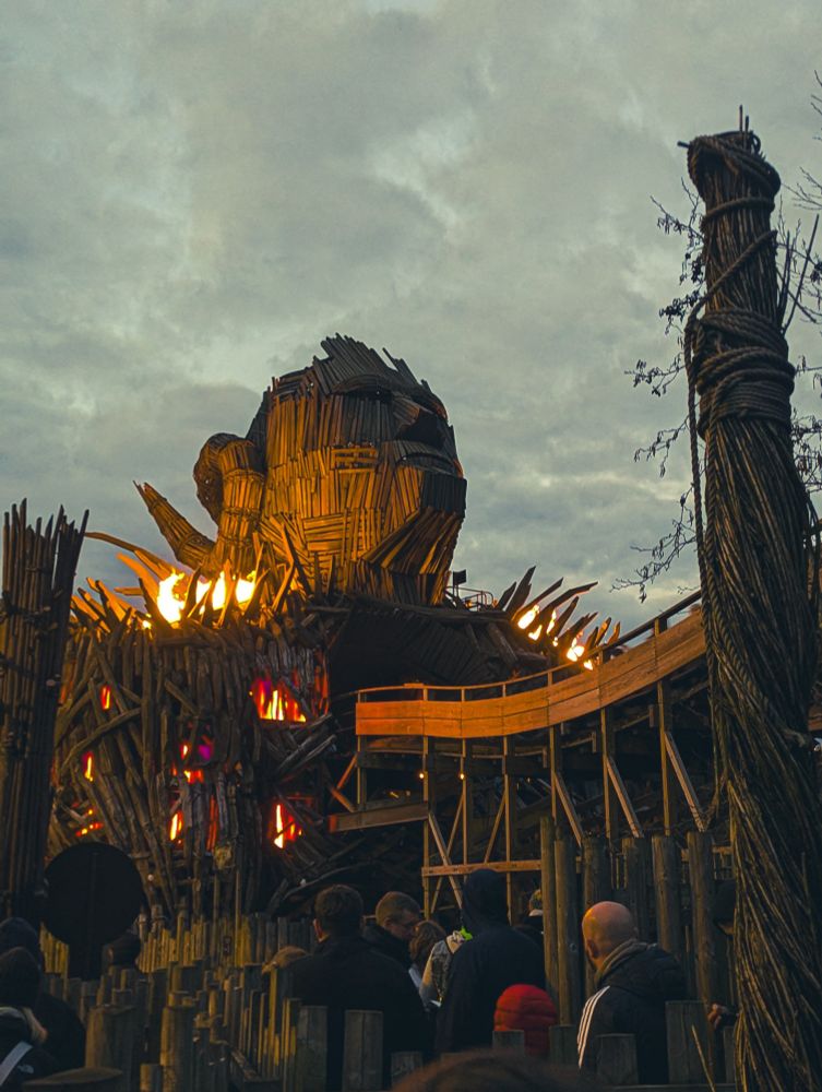 Wicker Man ride at Alton Towers at dusk from the queue.  The flames can be seen at either side of the wicker head structure. The rollercoaster is made of wood. 