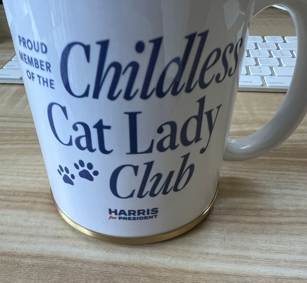 A mug with a brass ring around the base that is a perfectly fit coaster. The mug says Proud member of the childless cat lady club. Harris for President. 