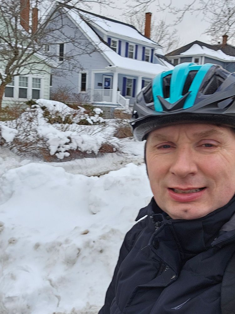 Me on a bike (which you can't see) with bike helmet, thick winter coat, with bick snow banks and houses in the background.