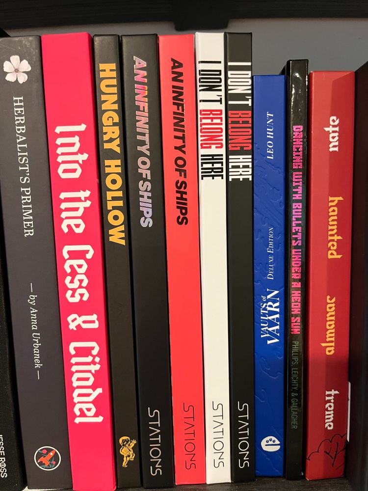 Side view of spines of books on a bookshelf. In the center of about eight books, two copies of An Infinity of Ships by STATIONS, and I Don't Belong Here by STATIONS