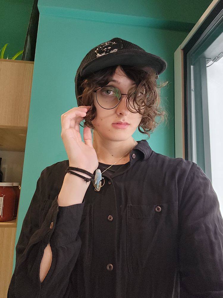 casval lour in a black baseball cap with a black beanie over it against the teal work wall wearing a black button up , she's got her hair over one of her eyes and she's wearing glasses (rare!)