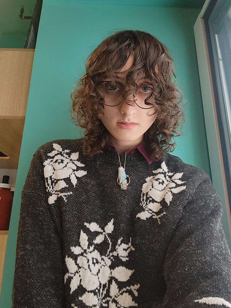 banagher lour against a teal wall in her black and white rose sweater with her red corduroy peaking out overtop with really cute hair too