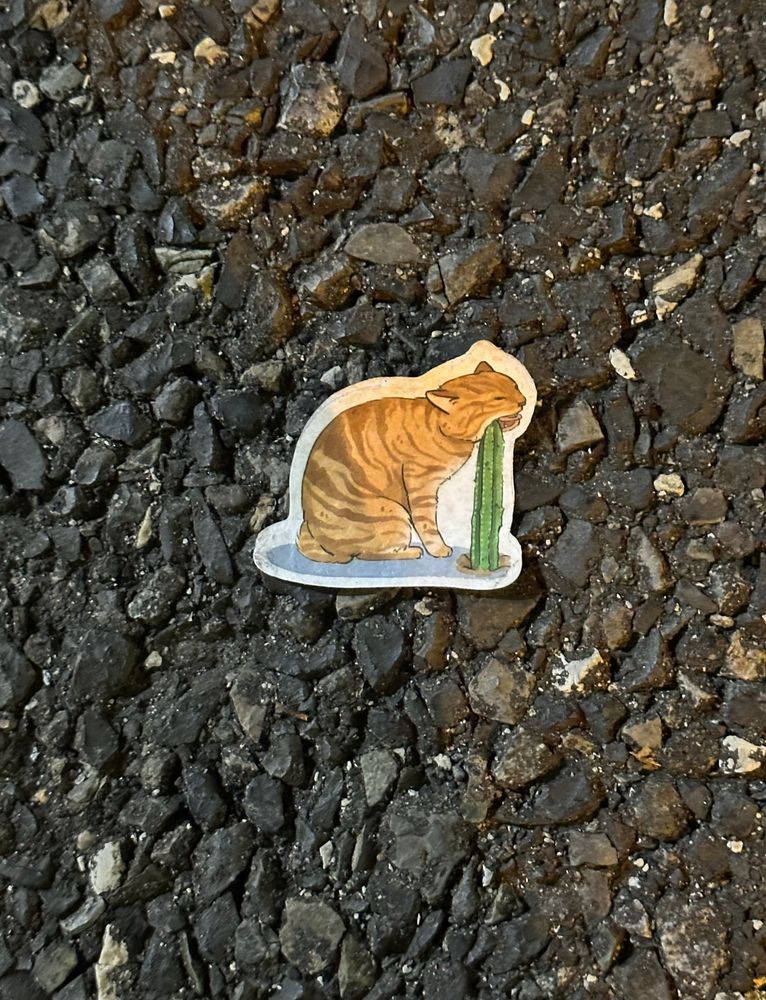 A third cat sticker on the ground, an orange cat nibbling on the top of a tall cactus. He does not look very happy