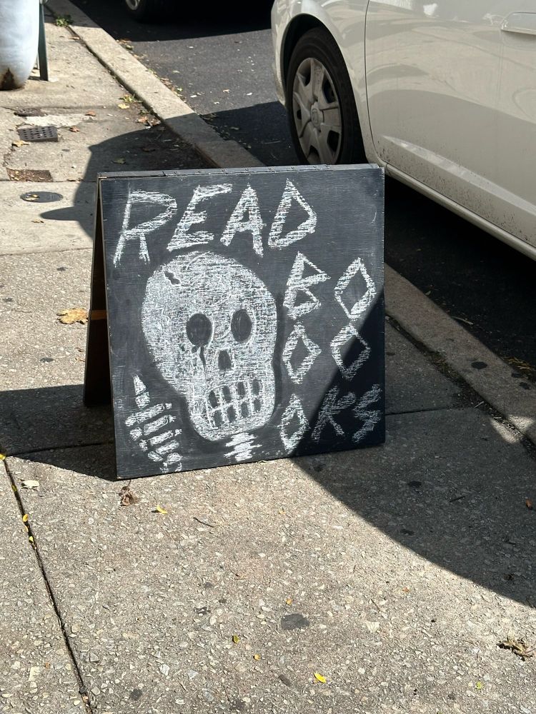 A friendly chalk drawn skeleton that is saying “read booooks”
