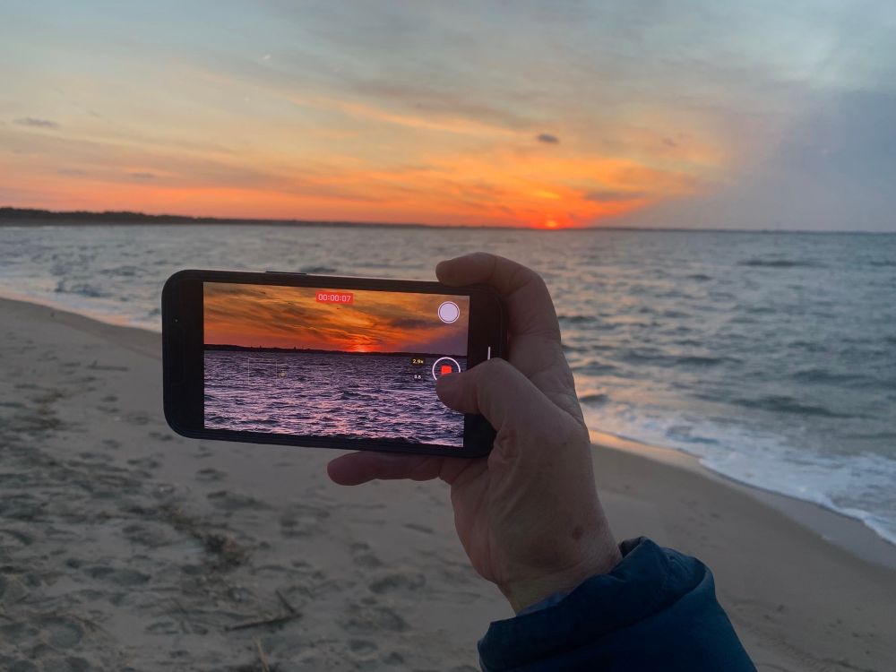 A gand holding an iPhone taking an image of the sunset in background 