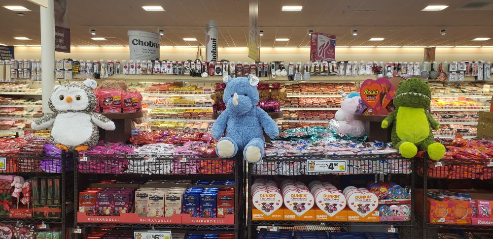 Atop a supermarket's long waist-high display of Valentine's candy sit three very large stuffed animals, each of them two or three feet tall.  There's an owl, a cute blue-grey rhino, and a stuffed olive and grass green t-rex.  The display is stacked to the gills and is very tidy and appealing.