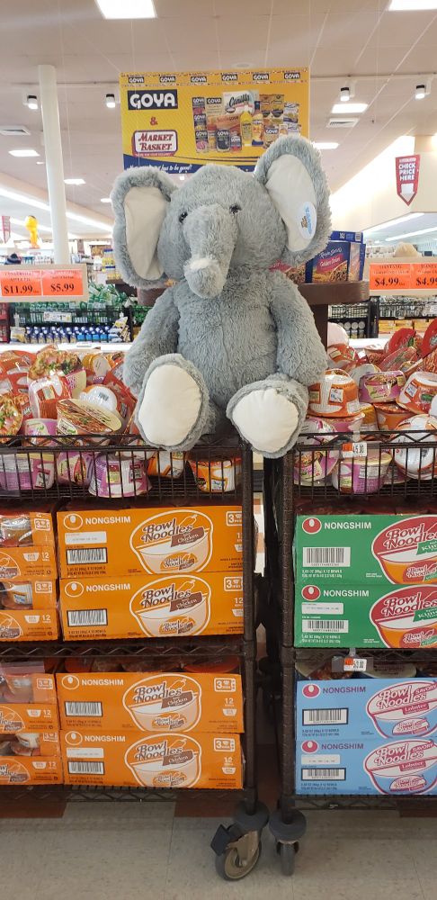 A giant 3 foot tall soft stuffed elephant sits in top of a large floor display full of ramen and other dried instant noodles.