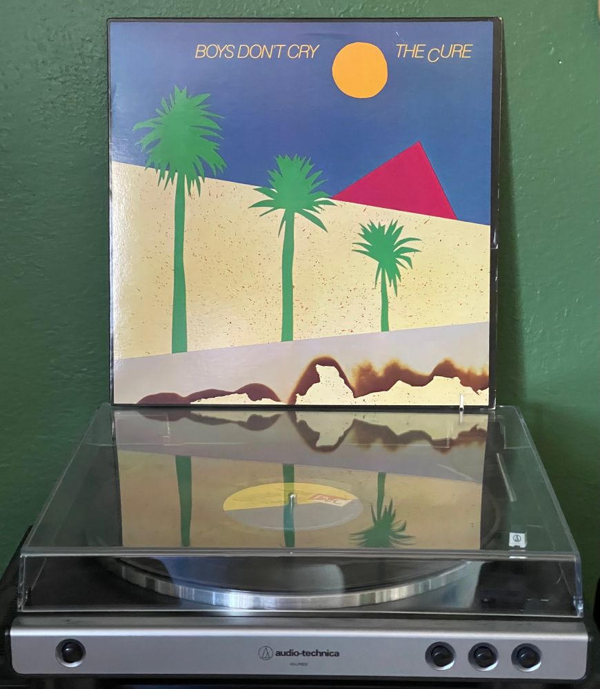 Turntable atop which is sitting a copy of The Cure “Boys Don’t Cry.”  Cover art is minimalist, depicting three palm trees in a desert, with a brilliant blue sky, and orange sun, and a pink pyramid in the background.