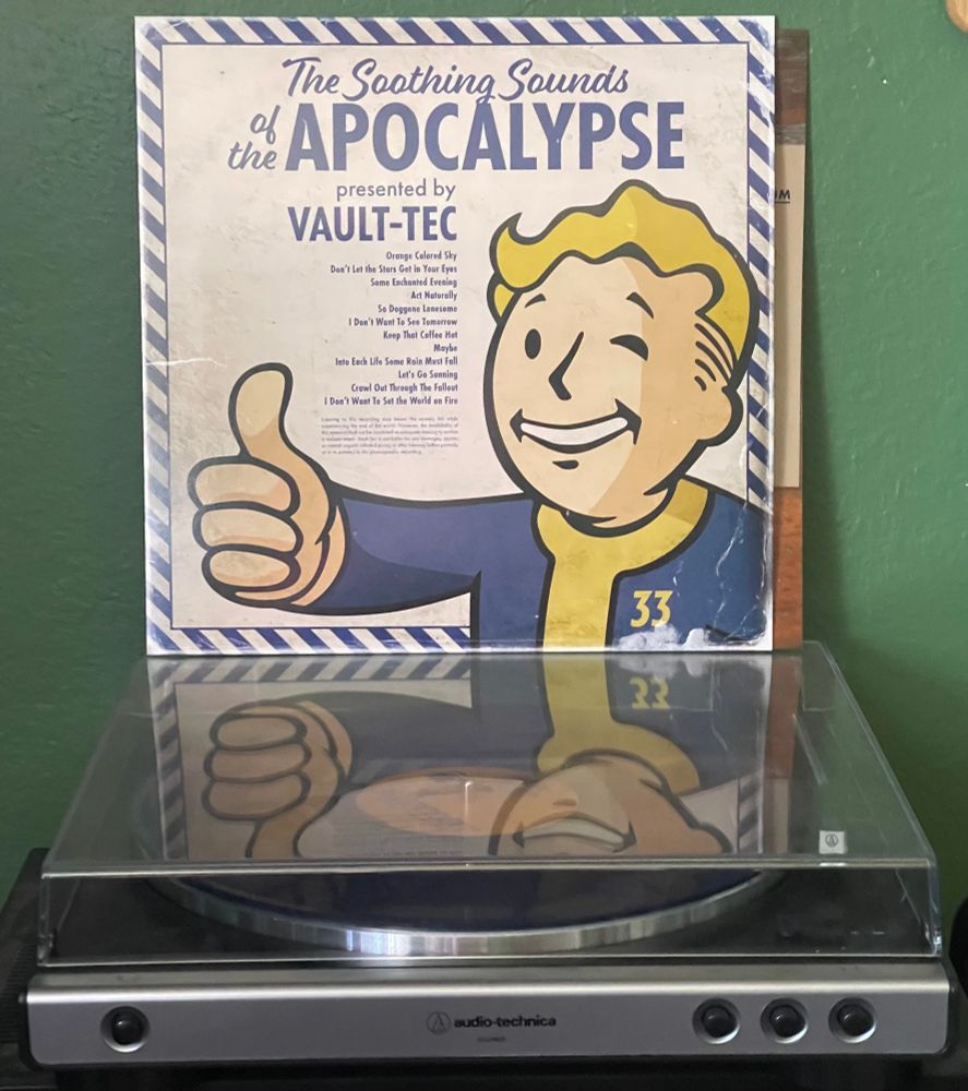 Copy of “The Sounds of the Apocalypse- Presented by Vault-Tec” perched atop a turntable. The cover art is MCM in styling, framing the Vault-Tec boy, giving a thumb’s up.