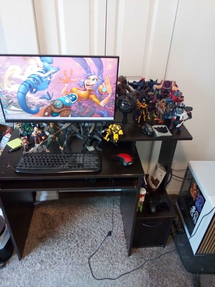 A computer desk with a PC tower with cartoony pinups of a xenomorph and yautja woman stickers on it, as well as a pair of stickers of Marina and Pearl from Splatoon. The PC monitor has a Wildstar background on it, and the desk is covered in Transformers toys, NECA Alien toys, a NECA Predator toy, a Play Arts toy of Agrias Oaks, a Wildstar pin, and a hand-made figurine of an OC.
