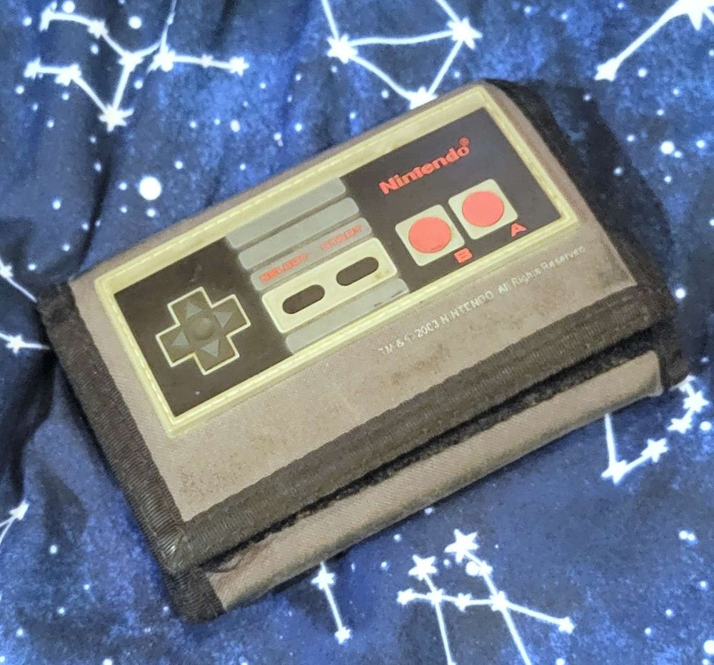 A gray velcro wallet with black trim, with a 3D NES controller decoration, laying on dark blue stars and constellations-themed bedsheets.