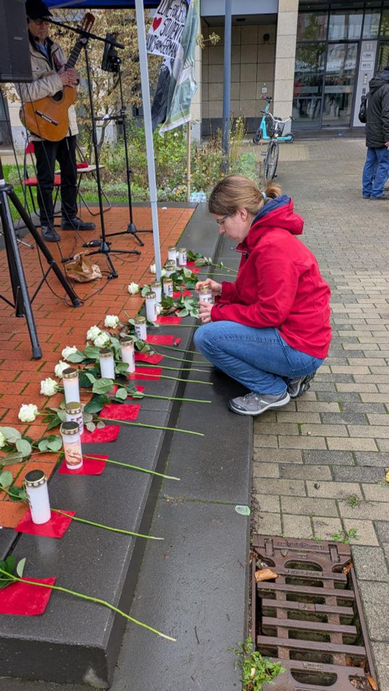 Ich hocke in einer roten Jacke vor einer Reihe Grabeskerzen mit weißen Rosen und zünde eine der Kerzen an. Dahinter sieht man eine Bühne aufgebaut.