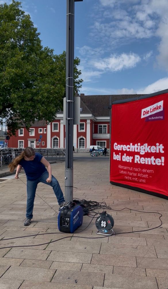 Eine Frau mit langen Haaren zieht an dem Starterseil eines Generators, während sie ihn mit dem Fuß festhält. Im Hintergrund steht ein Würfel mit dem Text "Gerechtigkeit bei der Rente!"