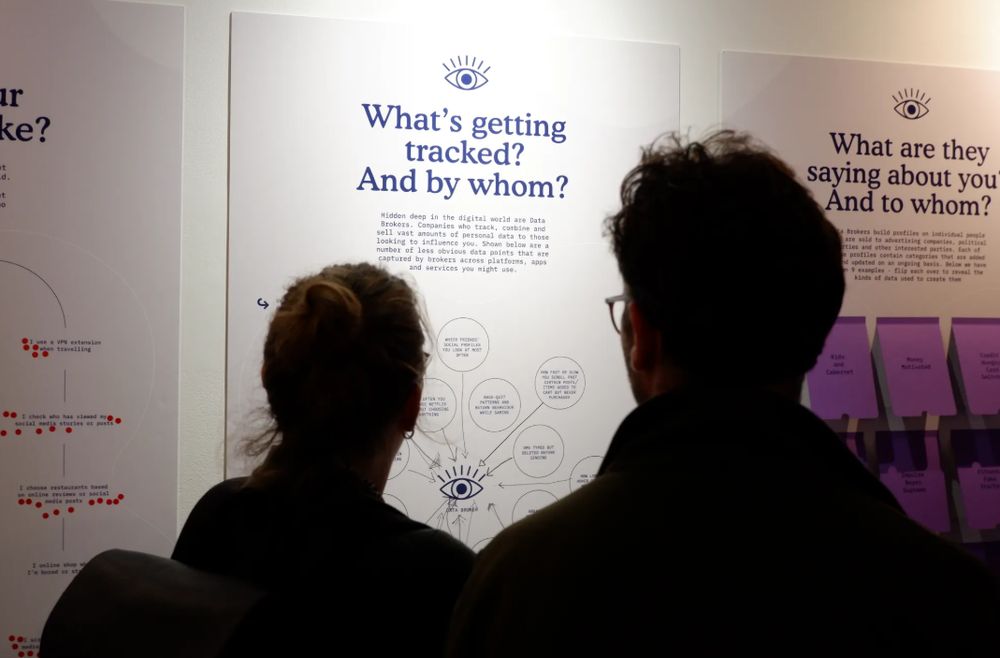 Two people, seen from behind, in silhouette, stand looking at a poster saying "Who's getting tracked? And by whom?" that is part of an exhibition on data brokers. 
