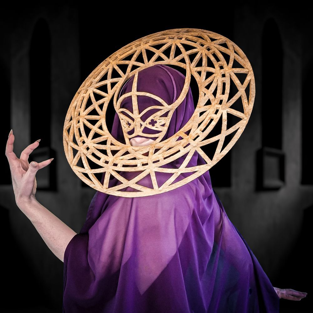 A wraith-like woman wearing a purple veil, a wine-red dress, and a gold necklace is standing in a dark chamber. The veil conceals her eyes, but she is looking towards the camera. Over the veil, the woman wears a carved sandstone headpiece comprising a half-mask with front and rear projections that support a ring torus encircling her head. The mask and the ring feature geometric cutouts referencing the camouflage pattern of a Gaboon viper and the crossed ceiling supports found in church vaults. The shape of the mask, with its slightly upturned nose bridge, resembles a snake’s head.