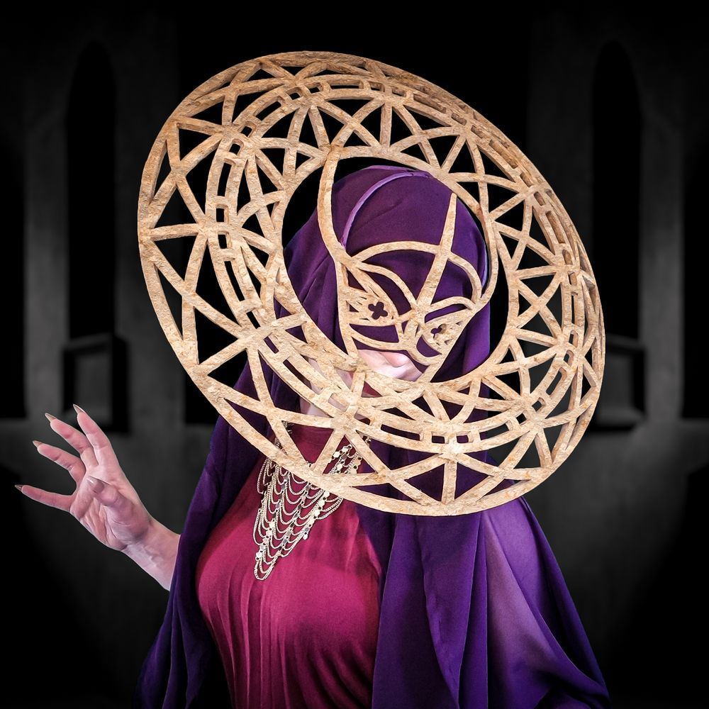 A wraith-like woman wearing a purple veil, a wine-red dress, and a gold necklace is standing in a dark chamber. The veil conceals her eyes, but she is looking towards the camera. Over the veil, the woman wears a carved sandstone headpiece comprising a half-mask with front and rear projections that support a ring torus encircling her head. The mask and the ring feature geometric cutouts referencing the camouflage pattern of a Gaboon viper and the crossed ceiling supports found in church vaults. The shape of the mask, with its slightly upturned nose bridge, resembles a snake’s head.